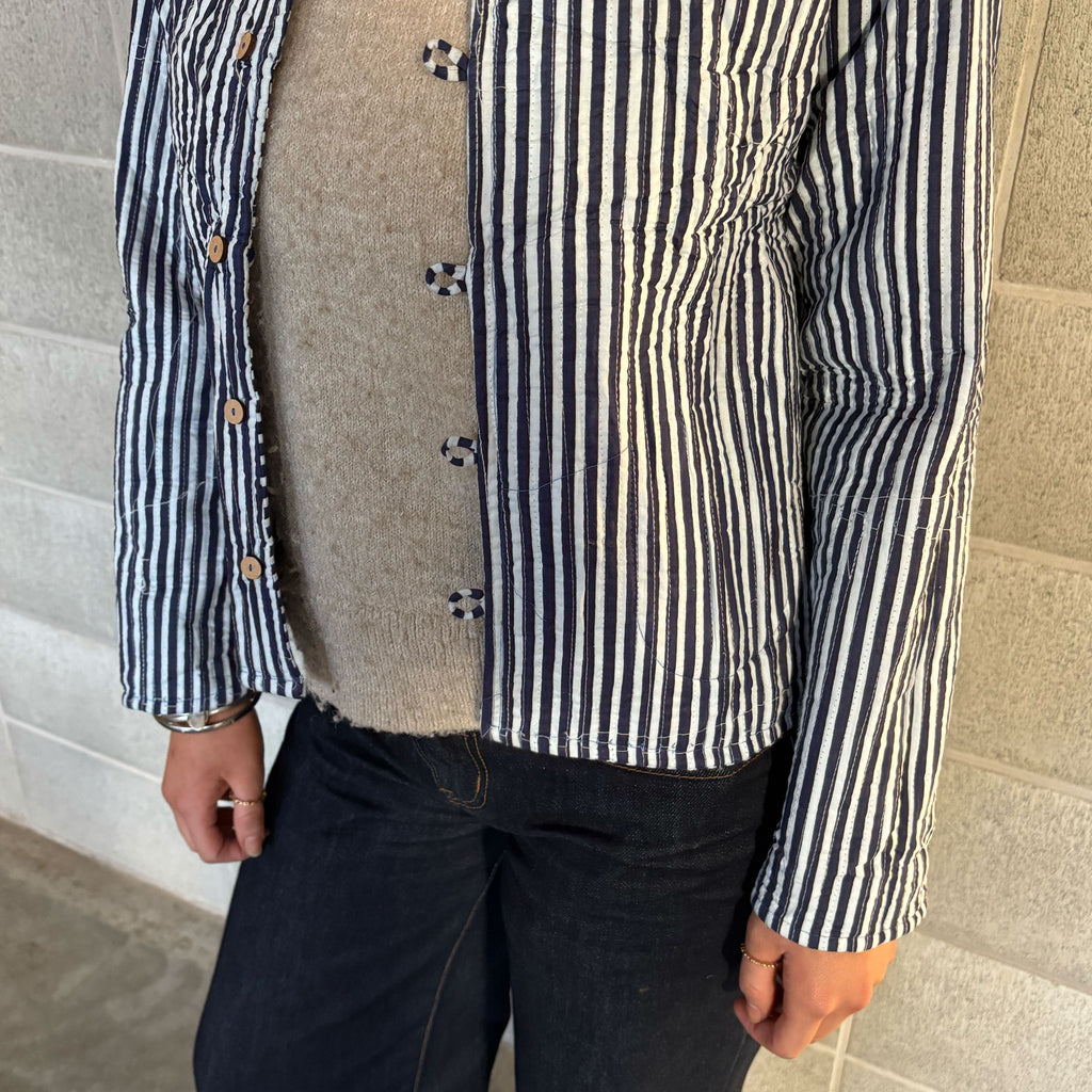 A person wearing a Colourstory navy-and-off-white vertically striped button-up jacket over a beige knit sweater and dark jeans, standing against a light stone wall.