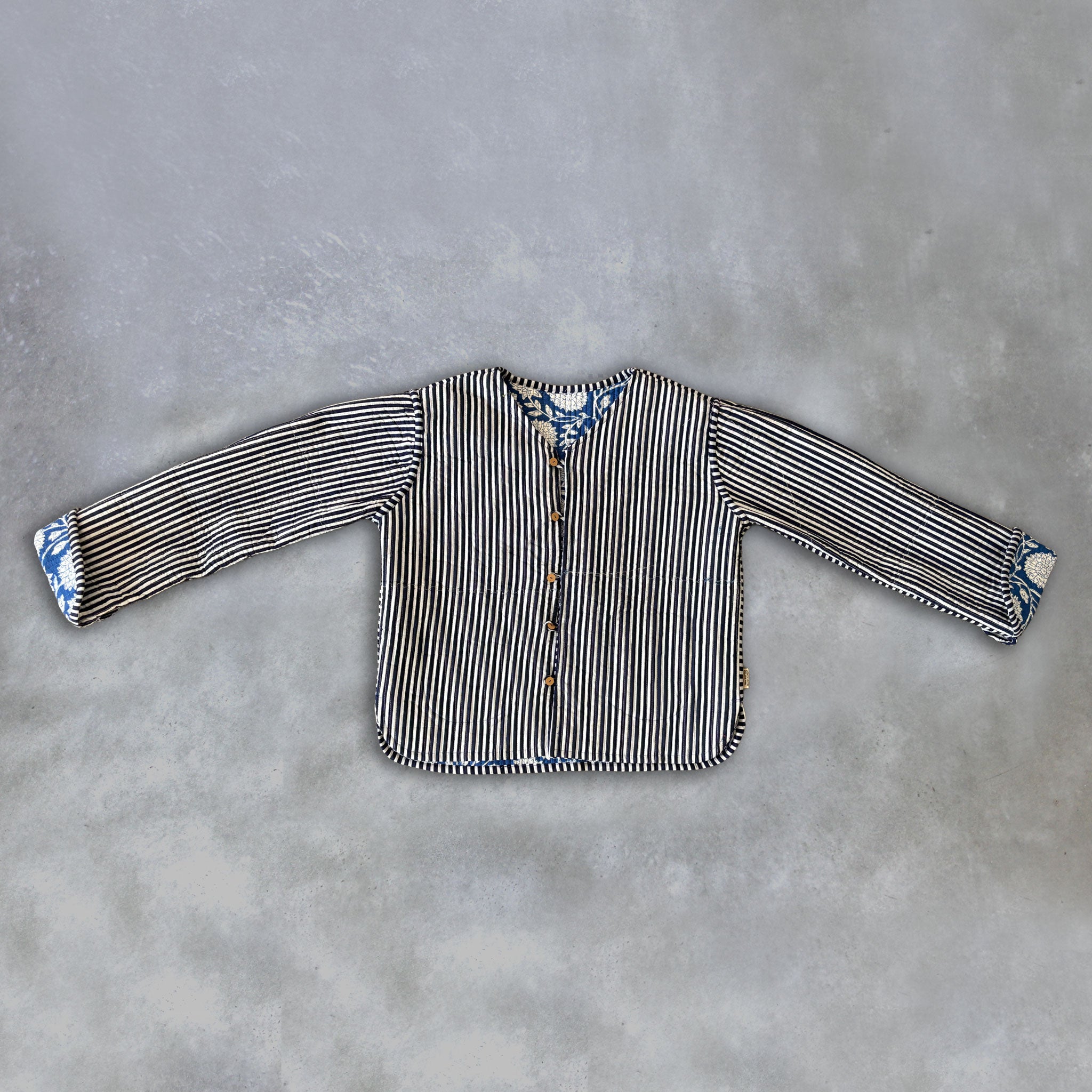 A long-sleeve inside of a Colourstory blue jacket striped with black and white vertical stripes and blue floral-patterned cuffs, laid flat on a textured grey background.