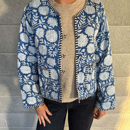 A person wearing a Colourstory, blue and white floral patterned quilted jacket over a beige sweater and dark jeans, standing against a gray brick wall.