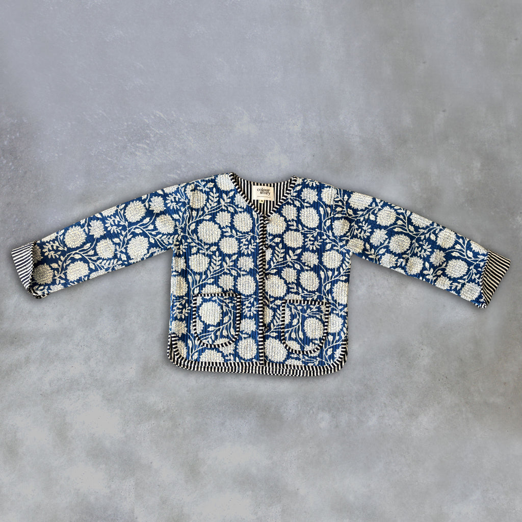 A long-sleeve quilted jacket by Colourstory featuring a blue and white floral block print with striped black and white trim, laid flat on a textured gray background.