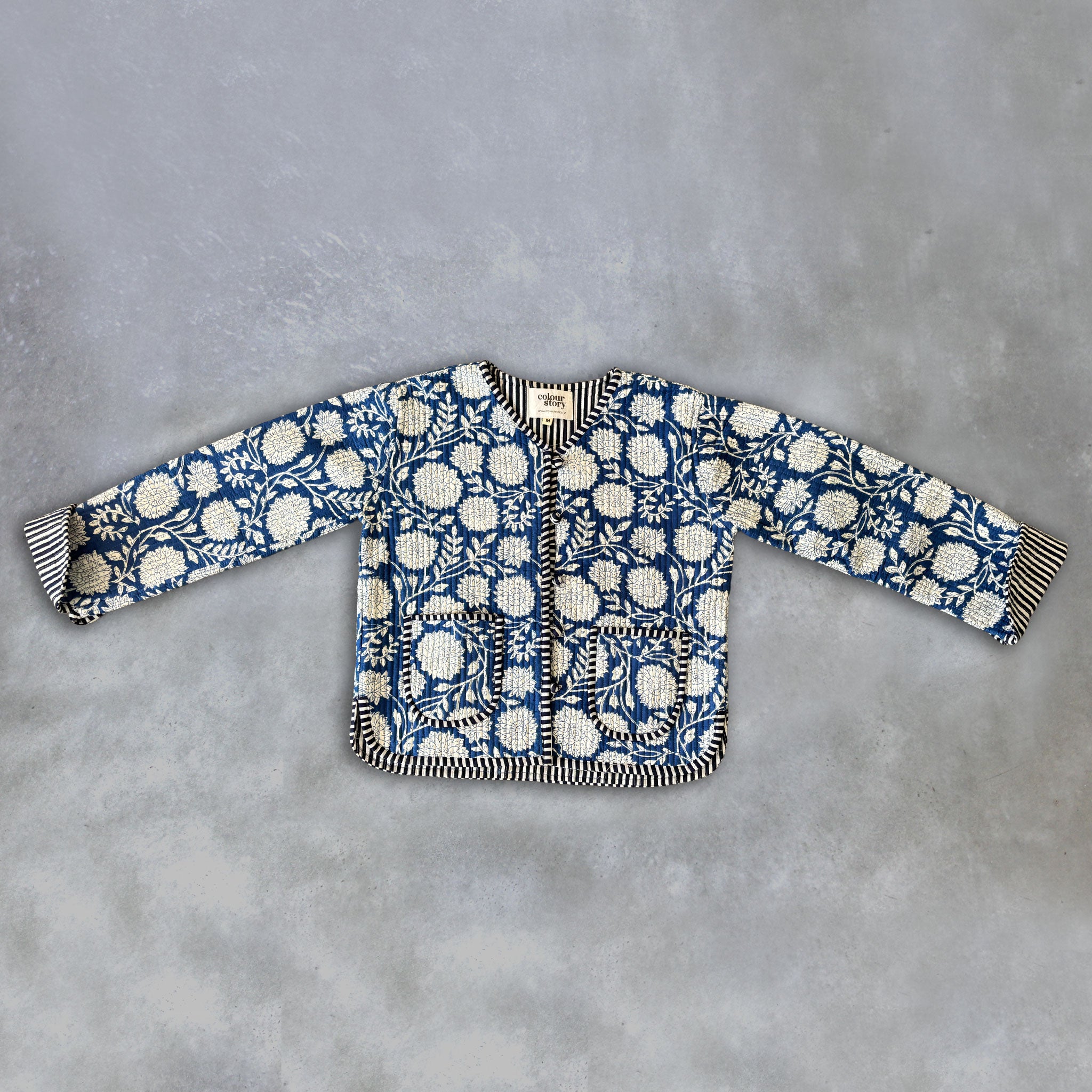 A long-sleeve quilted jacket by Colourstory featuring a blue and white floral block print with striped black and white trim, laid flat on a textured gray background.