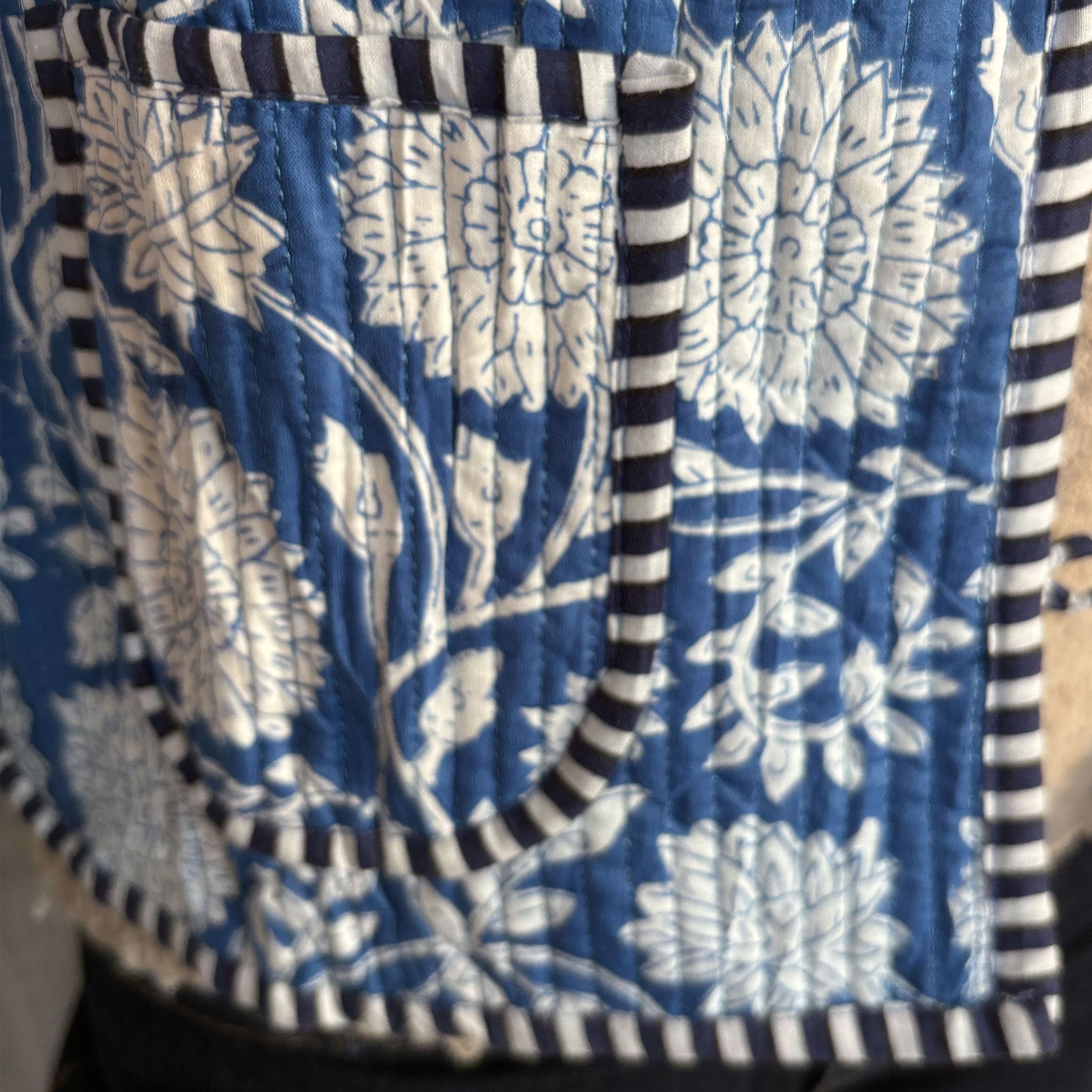 A close-up of a quilted Colourstory jacket,  featuring a blue and white floral block-print style pattern with dark navy and white striped piping on the pocket.