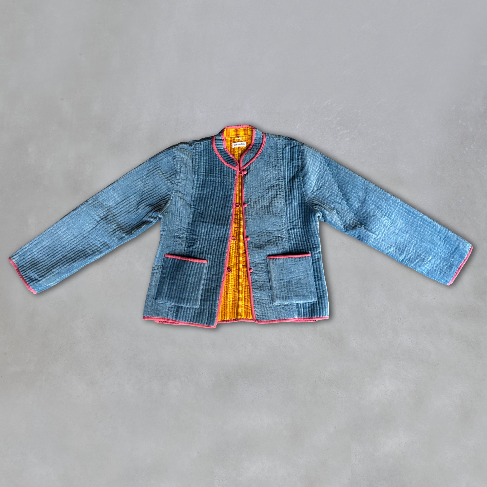 A blue velvet quilted jacket with a yellow and mustard stripe inner lining and pink piping trim, laid flat on a grey background.