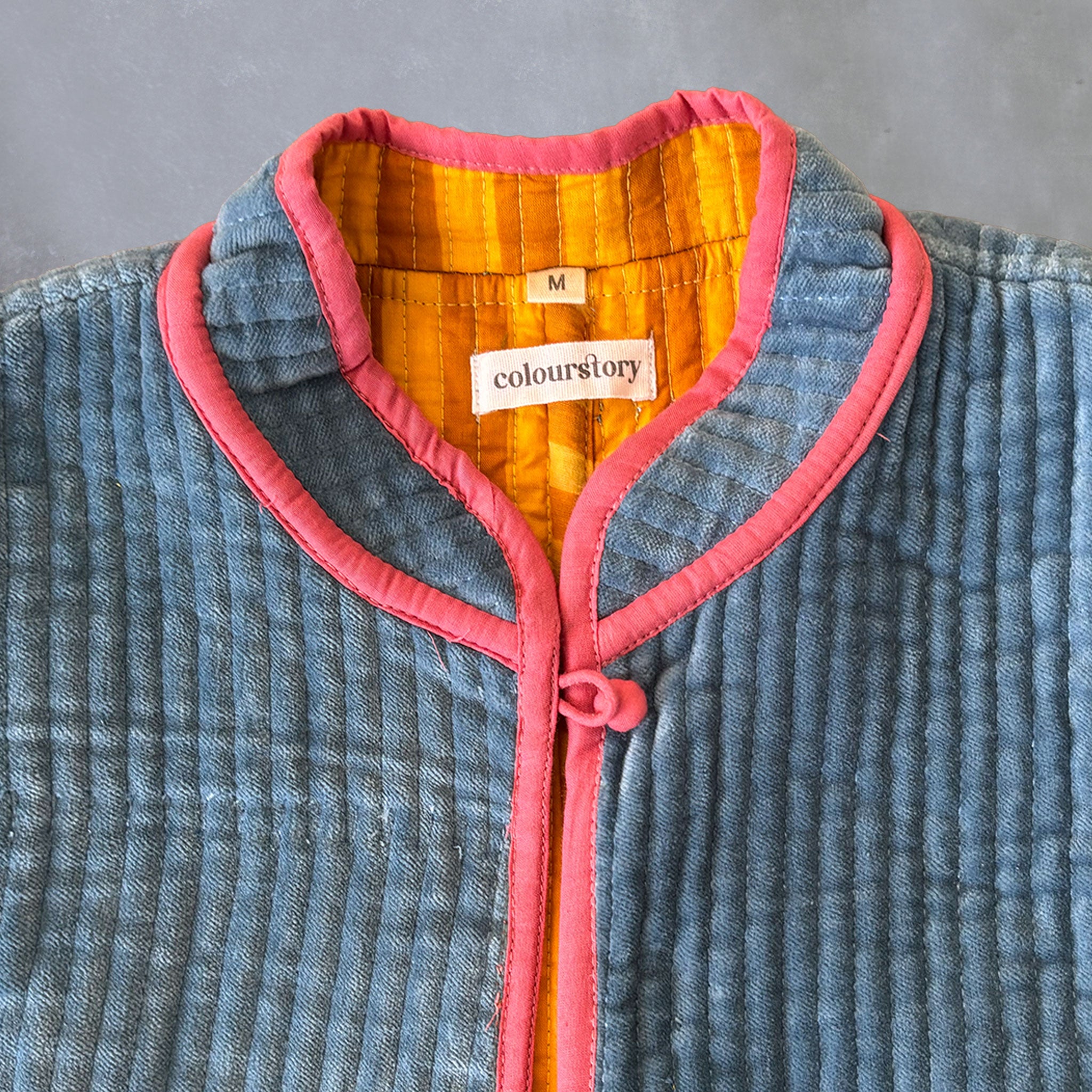 Close-up of a quilted dusty blue velvet textured jacket with a pink collar trim and button closure, showing a mustard and yellow striped inner lining and a 'Colourstory' brand label.