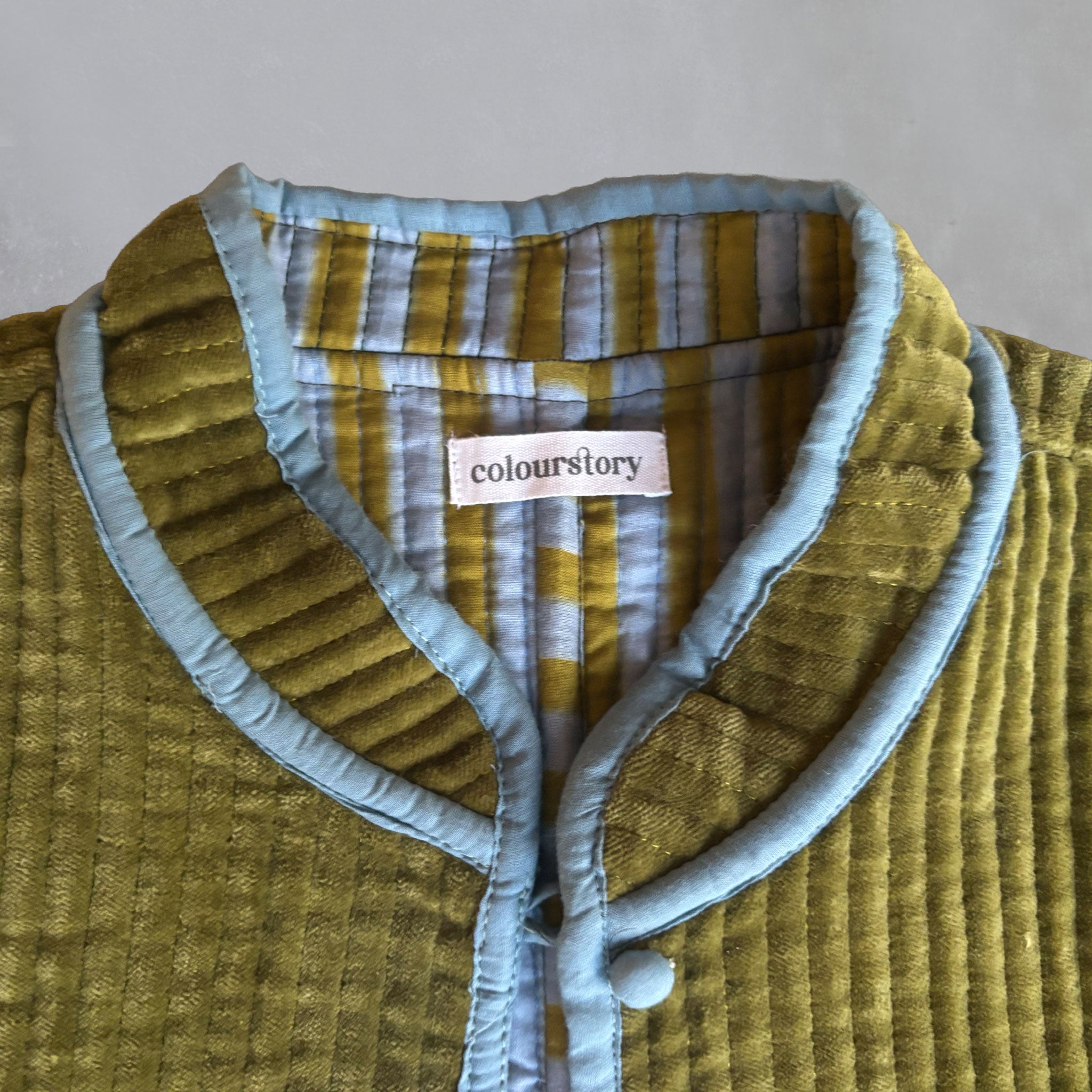 Close-up of a jacket with an olive velvet exterior, light blue piping, and a striped inner lining in light blue and olive. A visible neck label reads 'Colourstory'.