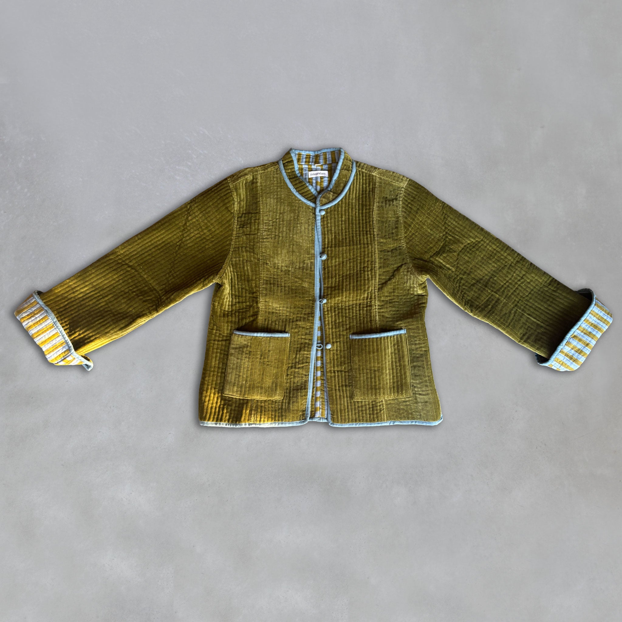 An olive green velvet jacket with light blue piping and striped olive-and-light blue cuff linings, laid flat on a grey background. Front pockets.