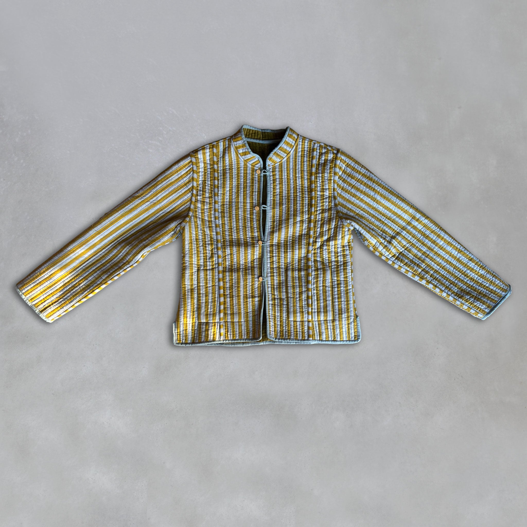 A long-sleeve Colourstory quilted jacket with a mandarin collar, featuring an olive and light blue striped pattern, laid flat on a textured grey background.