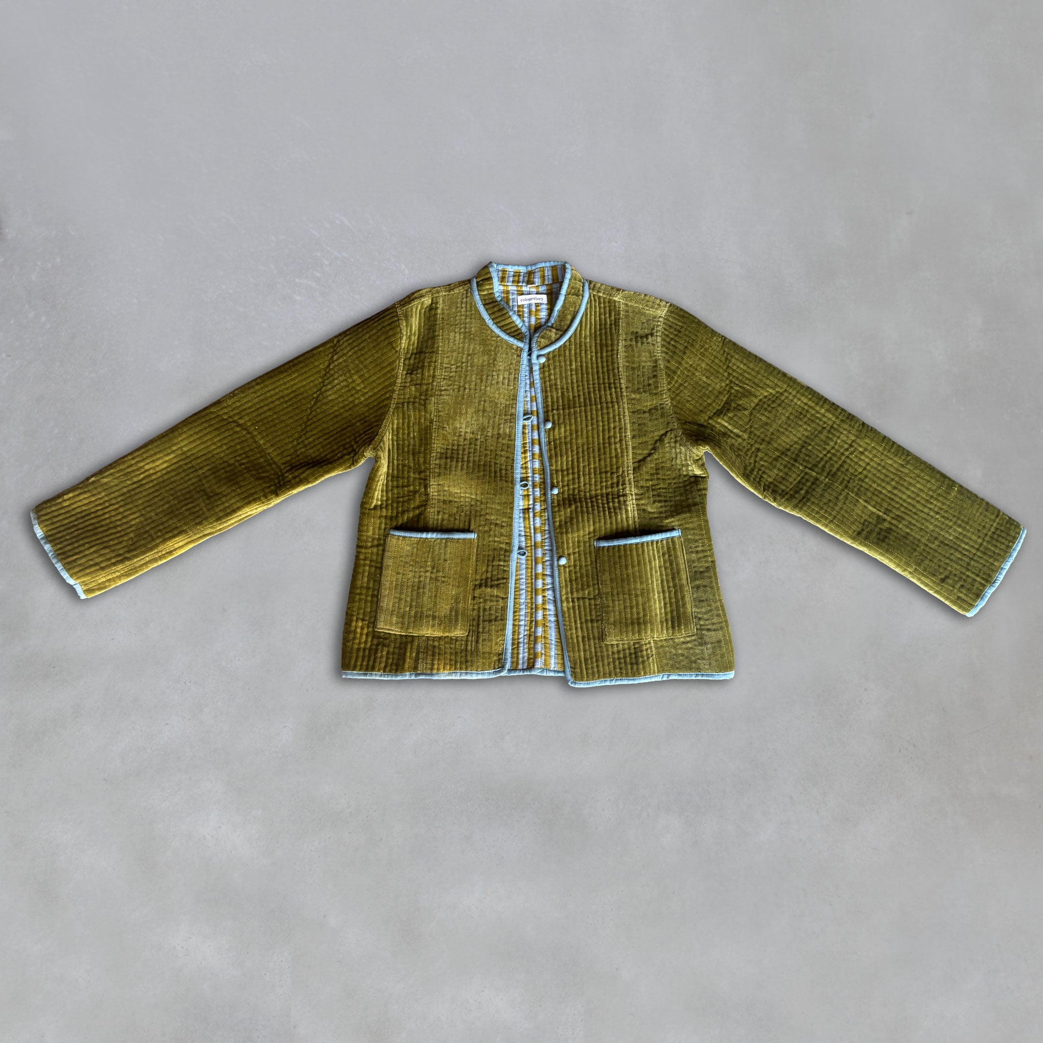 An olive green velvet jacket with light blue piping and a decorative button placket, laid flat on a grey background.