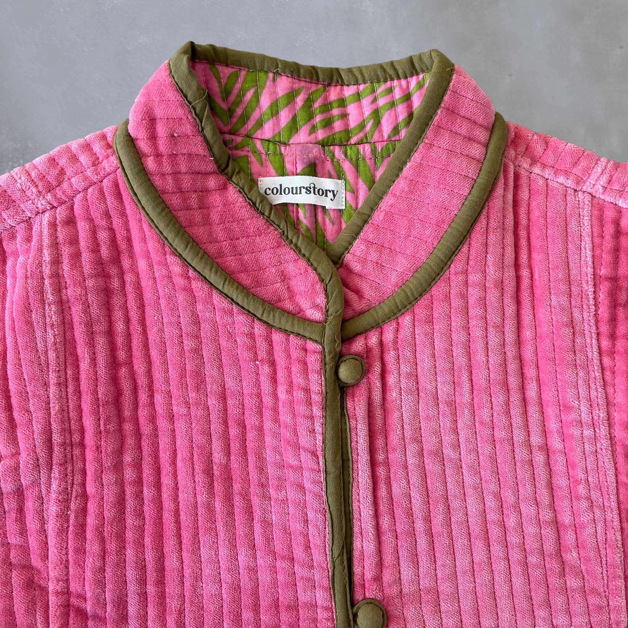 A close-up of a pink quilted velvet jacket with olive green trim and a green & pink patterned inner lining, featuring a visible Colourstory brand label.