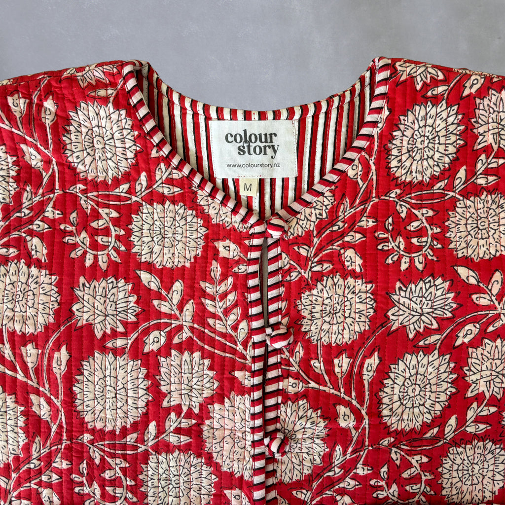 A close-up of quilted red cotton jacket with a cream floral block print, featuring a striped V-neck trim and a visible 'colour story' brand label.