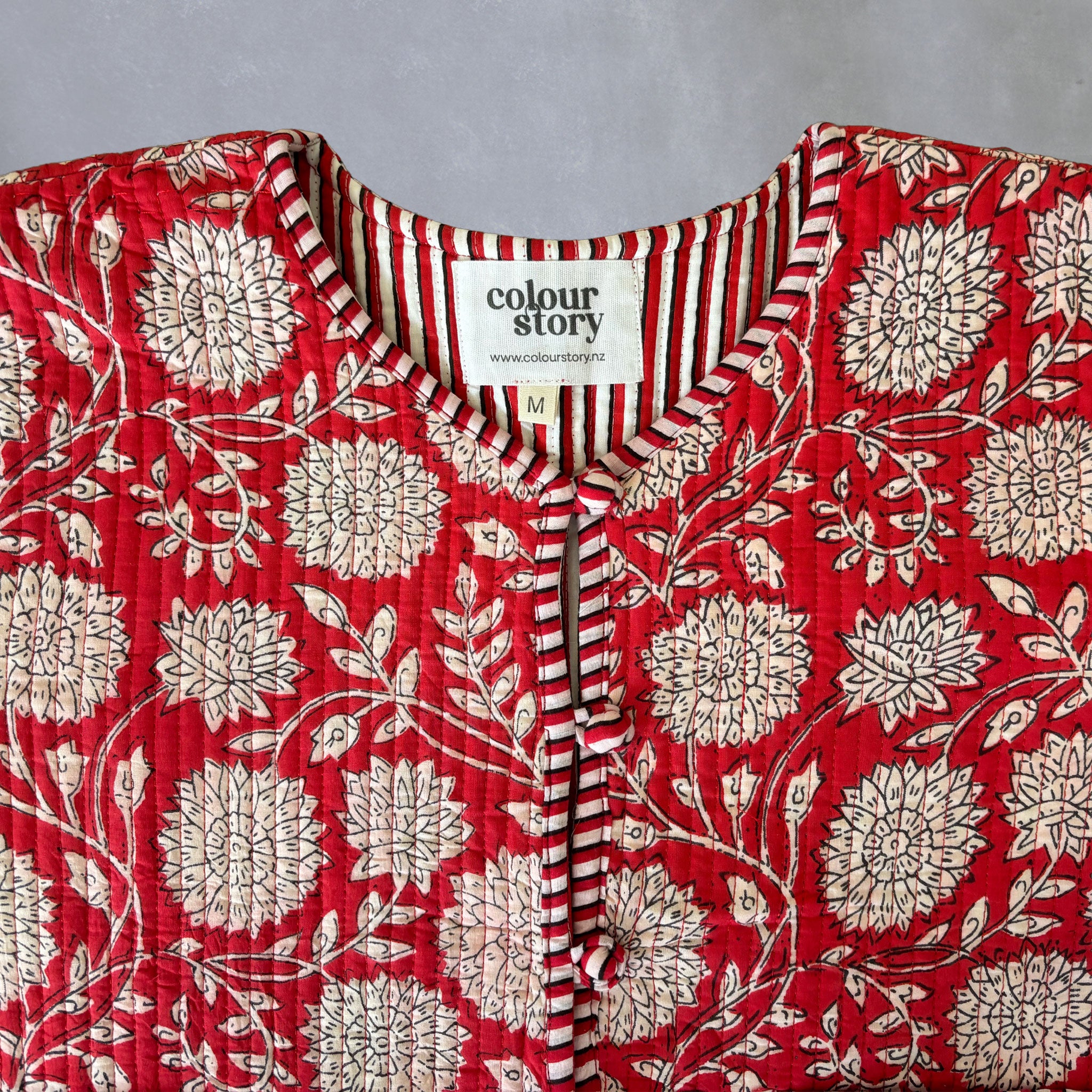 A close-up of quilted red cotton jacket with a cream floral block print, featuring a striped V-neck trim and a visible 'colour story' brand label.