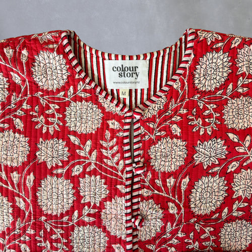 A close-up of quilted red cotton jacket with a cream floral block print, featuring a striped V-neck trim and a visible 'colour story' brand label.