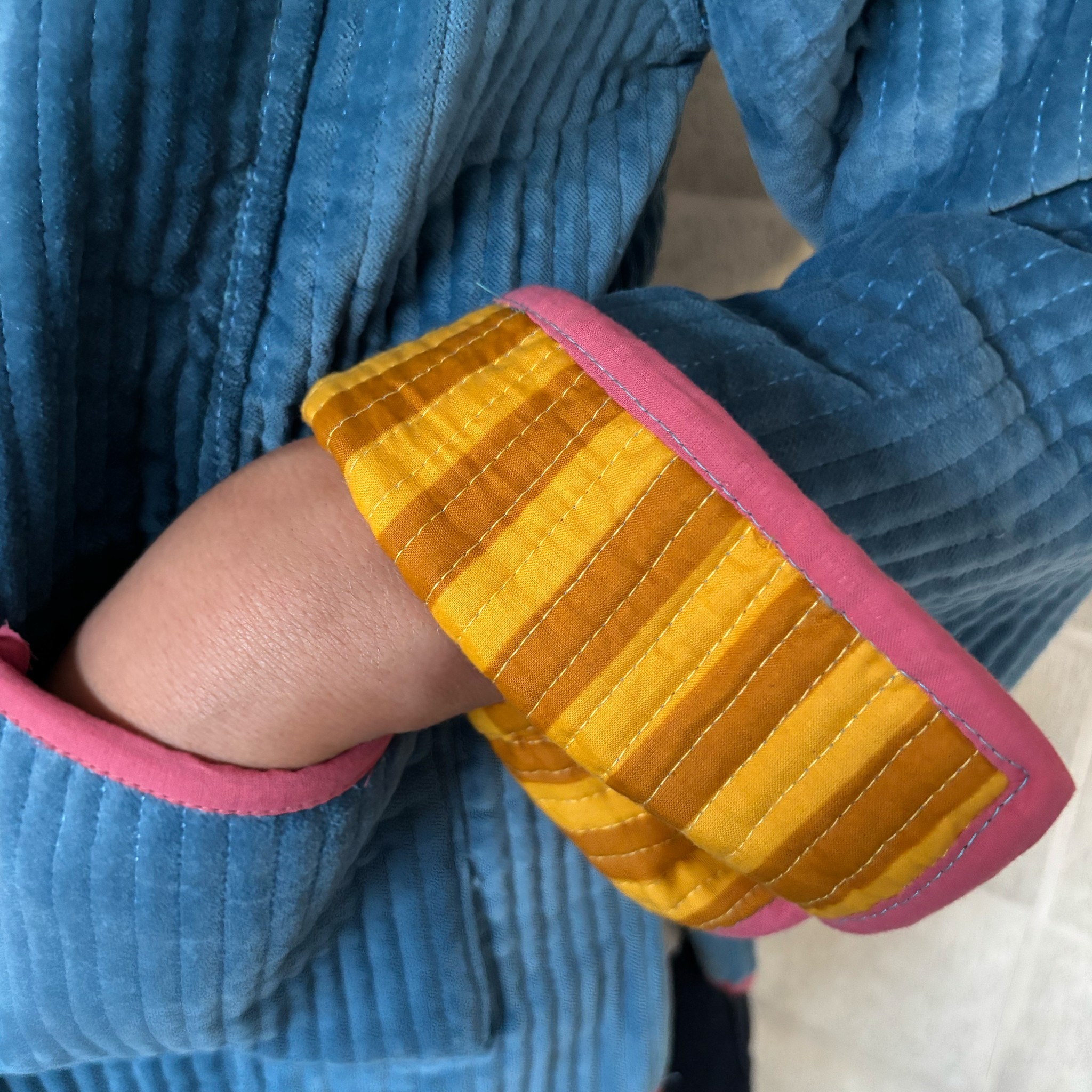 Close-up of a smokey blue velvet jacket with a folded cuff showing a yellow and mustard striped lining and pink trim, worn by a person.