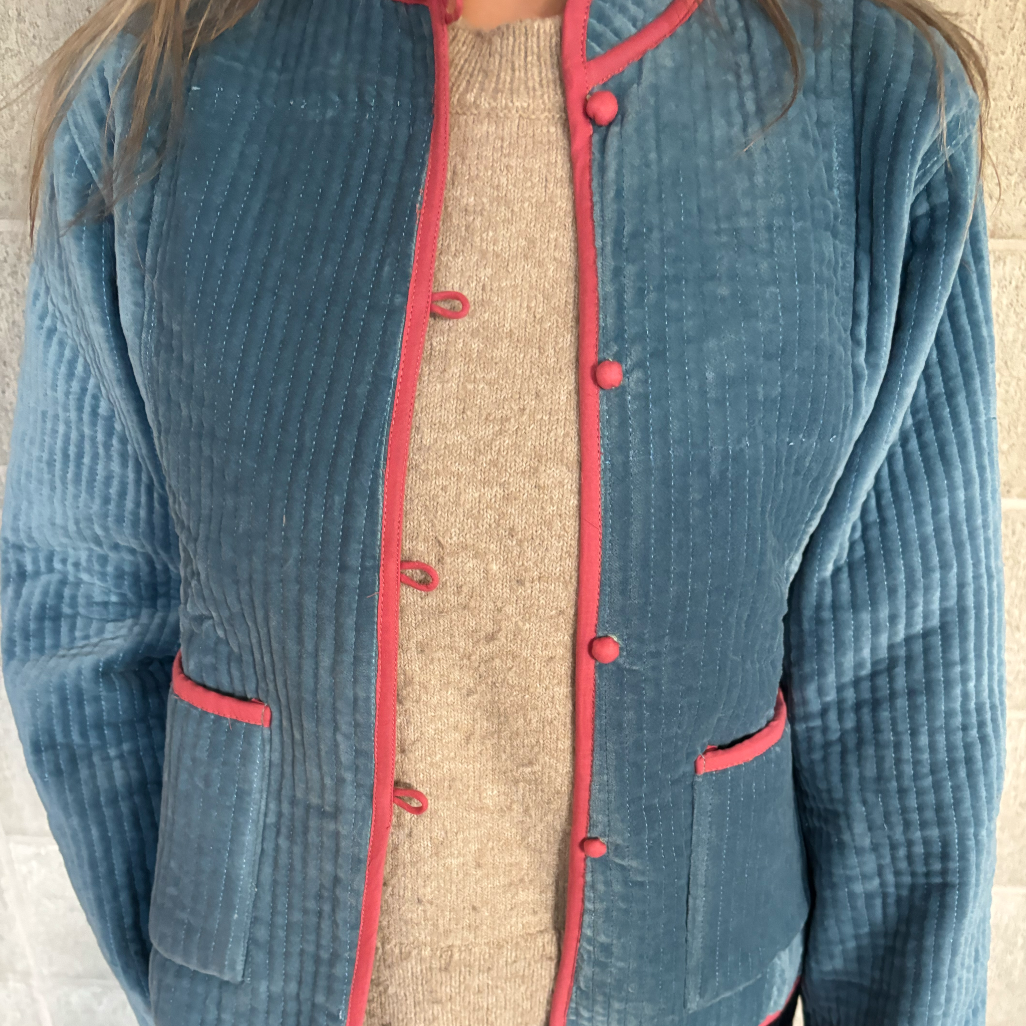 A close-up of a person wearing a smokey blue quilted velvet jacket with pink trim and matching buttons, layered over a beige knit sweater.