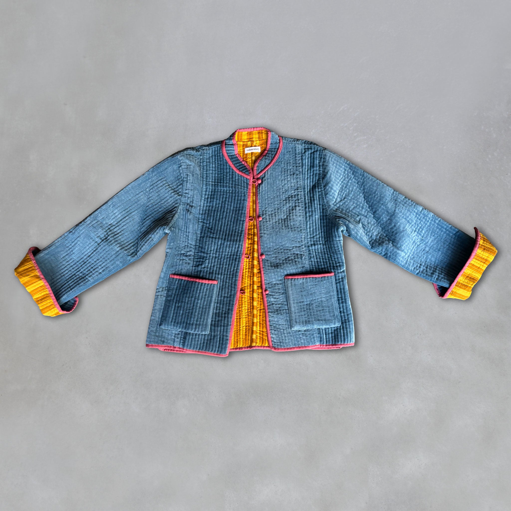 A blue quilted velvet jacket with yellow-and-mustard-striped cuffs and collar, and a yellow-striped, reversible inside jacket laid flat on a grey background.