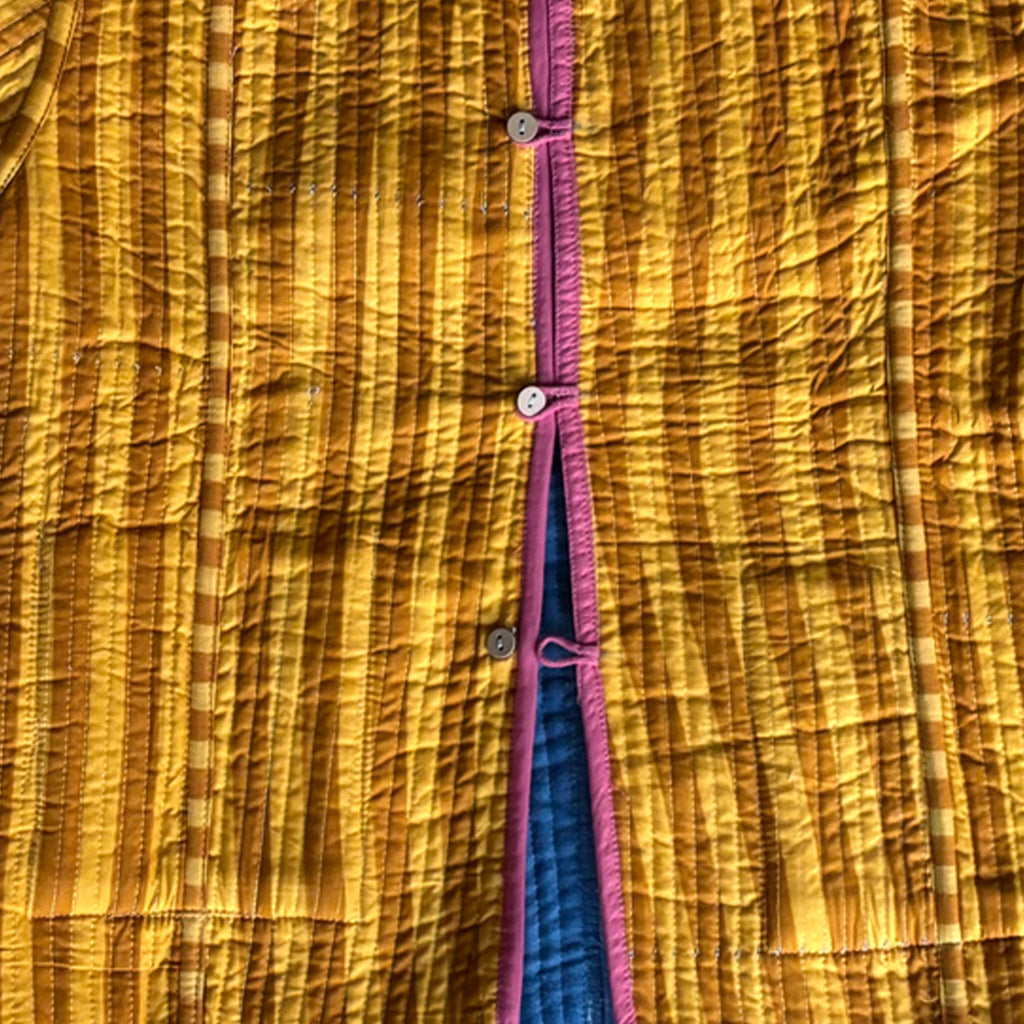 Close-up of a quilted jacket with mustard yellow stripes with a vertical pink trim and buttons with a dusty blue velvet visible along the opening.