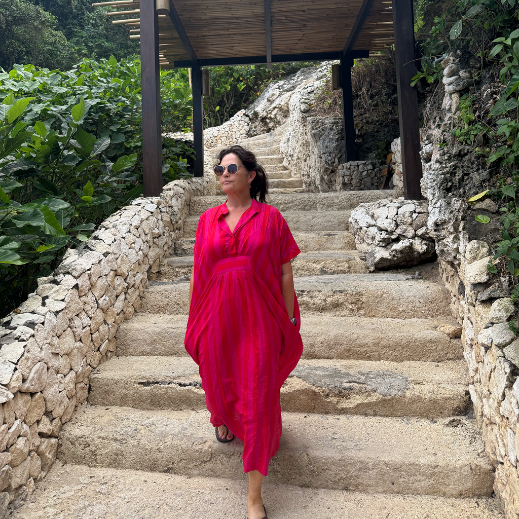 Tracey, wearing our bright pinkish-red kaftan maxi dress, walks down outdoor stone stairs under a wooden pavilion with lush green foliage on both sides.