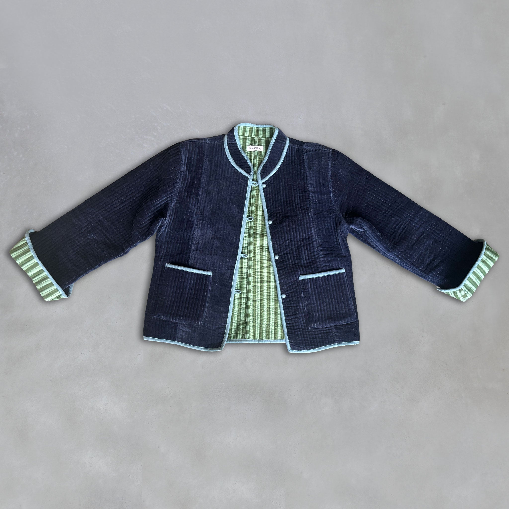 A dark navy velvet jacket with light blue piping and green-and-sage green striped cuffs, laid flat on a textured grey background.