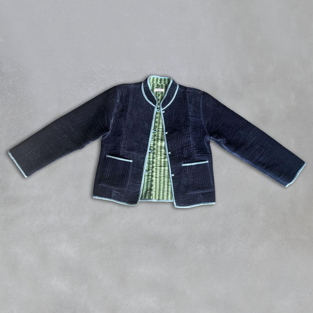 A dark navy velvet jacket with light blue piping, laid flat on a textured grey background. The jacket is open to reveal a green-and-sage-green striped lining.
