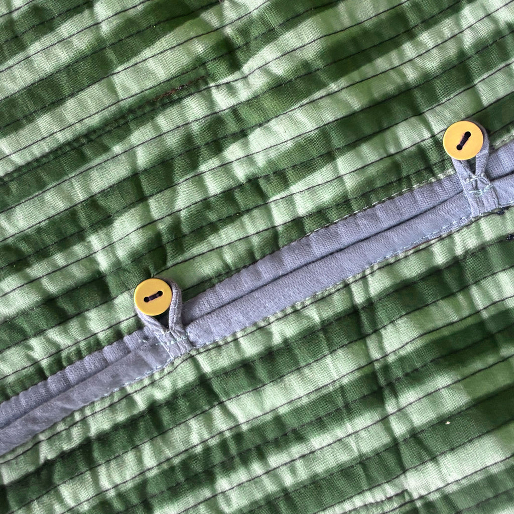 Close-up of a forest green and sage green striped fabric garment with a light blue trim and two buttons, showing the texture and stitching details.
