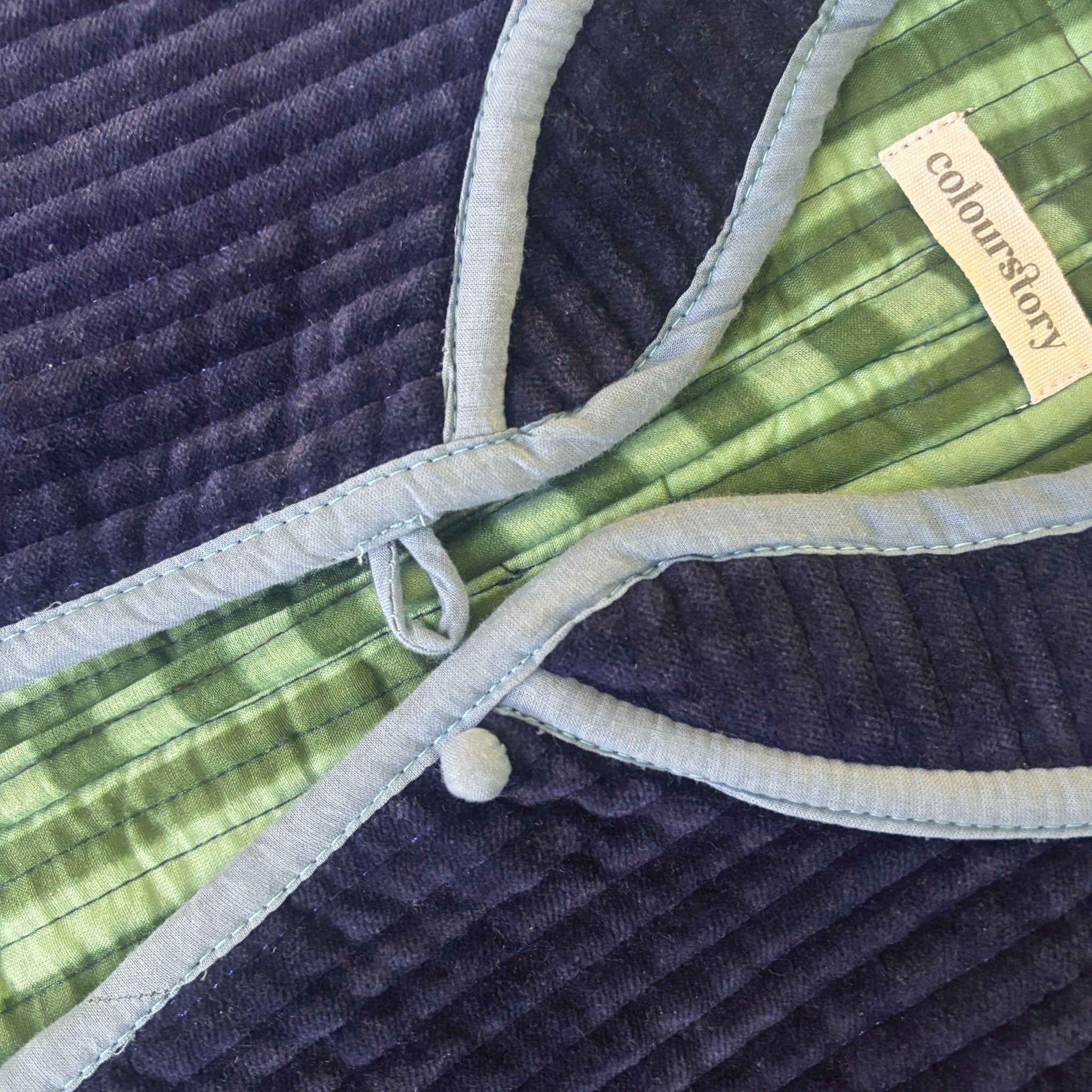 Close-up of a quilted Colourstory jacket showing dark navy velvet quilted fabric, a forest green and sage green striped inside, pale blue piping, and a small sewn-in label reading 'Colourstory'.