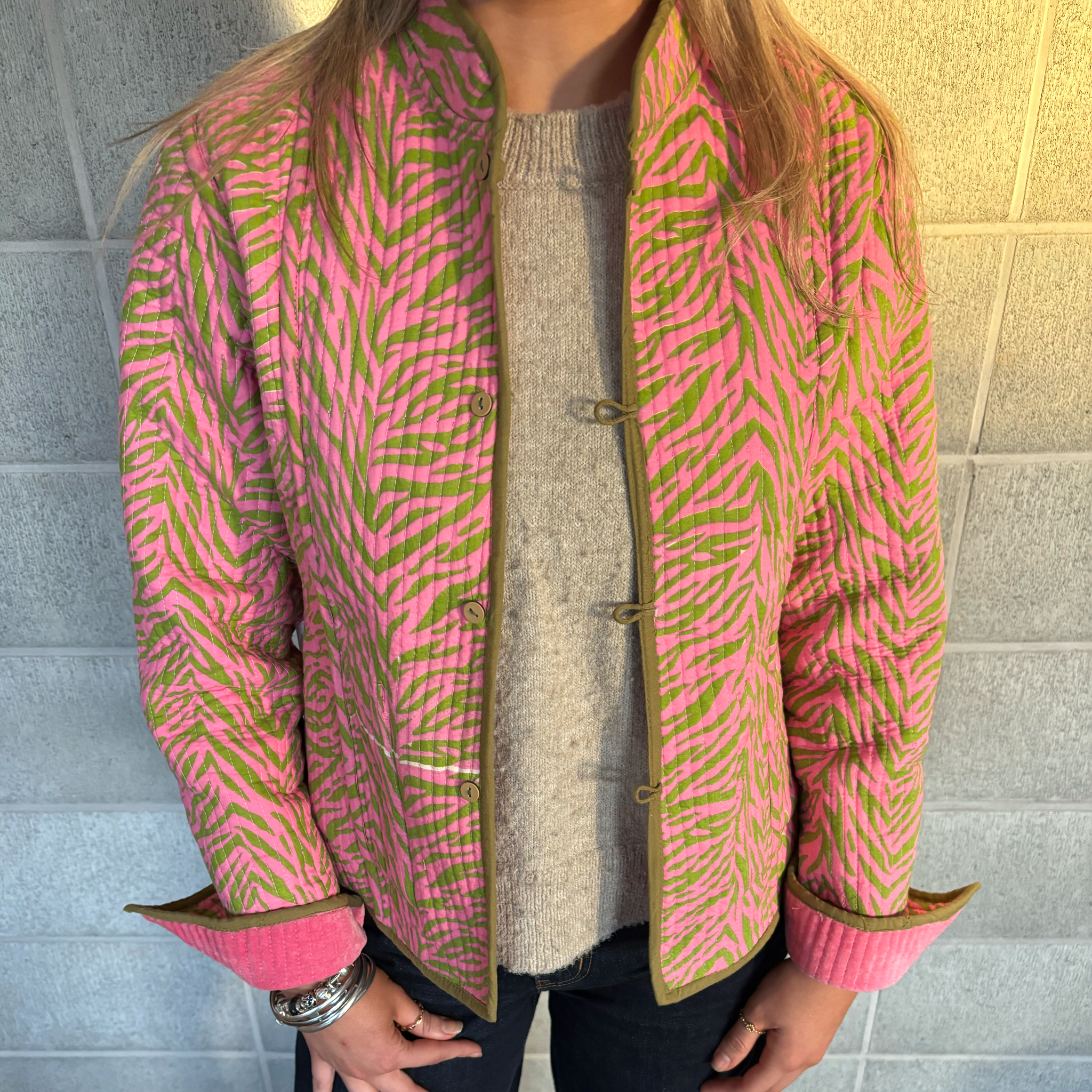 ColourStory model wearing a bright pink and green patterned jacket with olive green trim and patterned cuff lining, layered over a beige knit sweater, standing against a light stone wall.