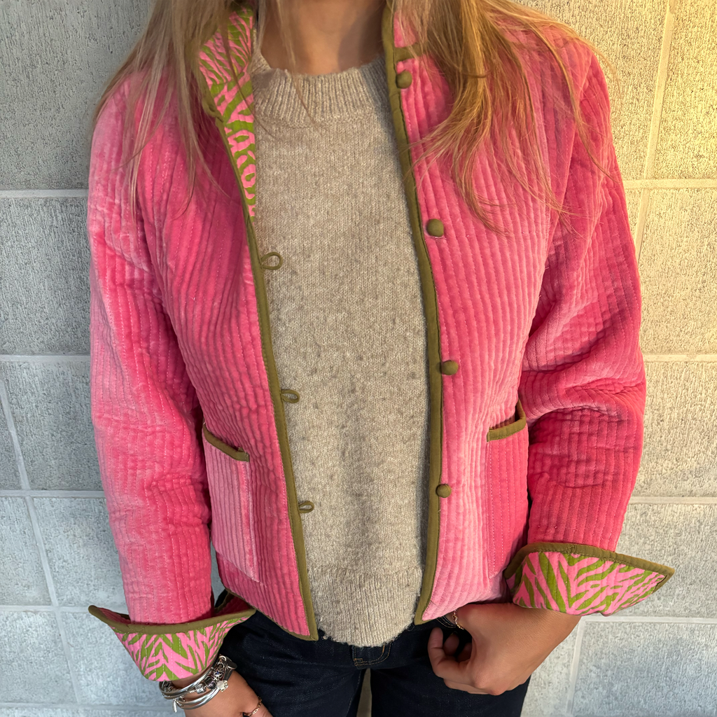 Colourstory model wearing a bright pink velvet jacket with olive green trim and patterned cuff lining, layered over a beige knit sweater, standing against a light stone wall.
