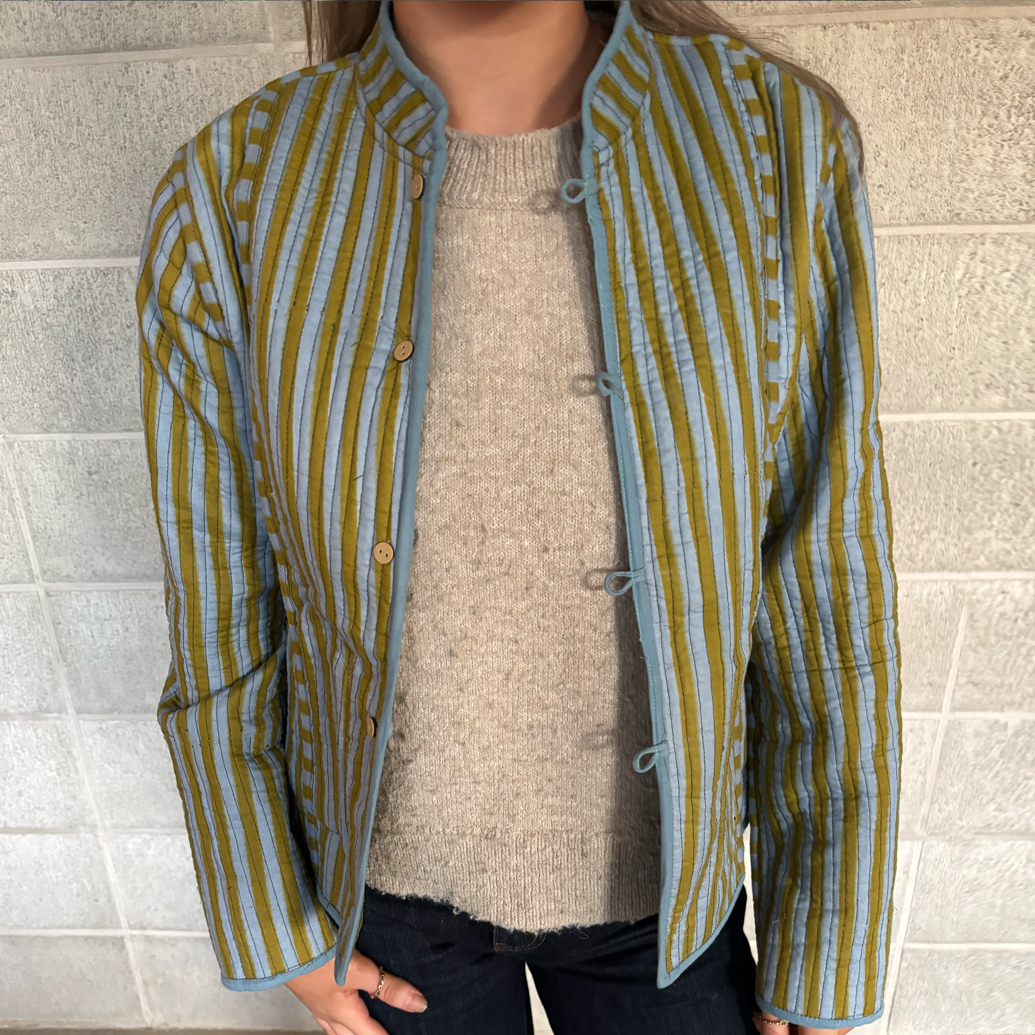 Our ColourStory model is wearing a cropped, long-sleeve quilted jacket with vertical olive green and light blue stripes, featuring a mandarin collar and button-tab closure, layered over a beige knit sweater.