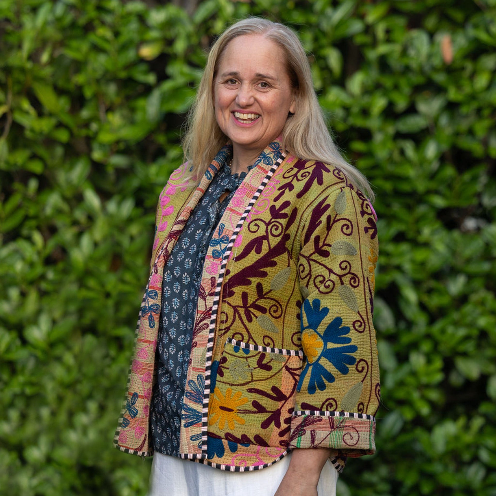Woman (Lucy) wearing a colourful embroidered jacket by Colourstory standing in front of green foliage.
