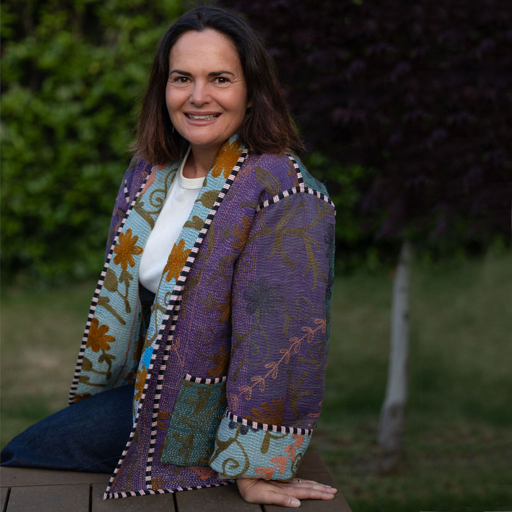 Woman (Tracey) wearing a colourful, patterned jacket sitting outdoors with greenery in the background