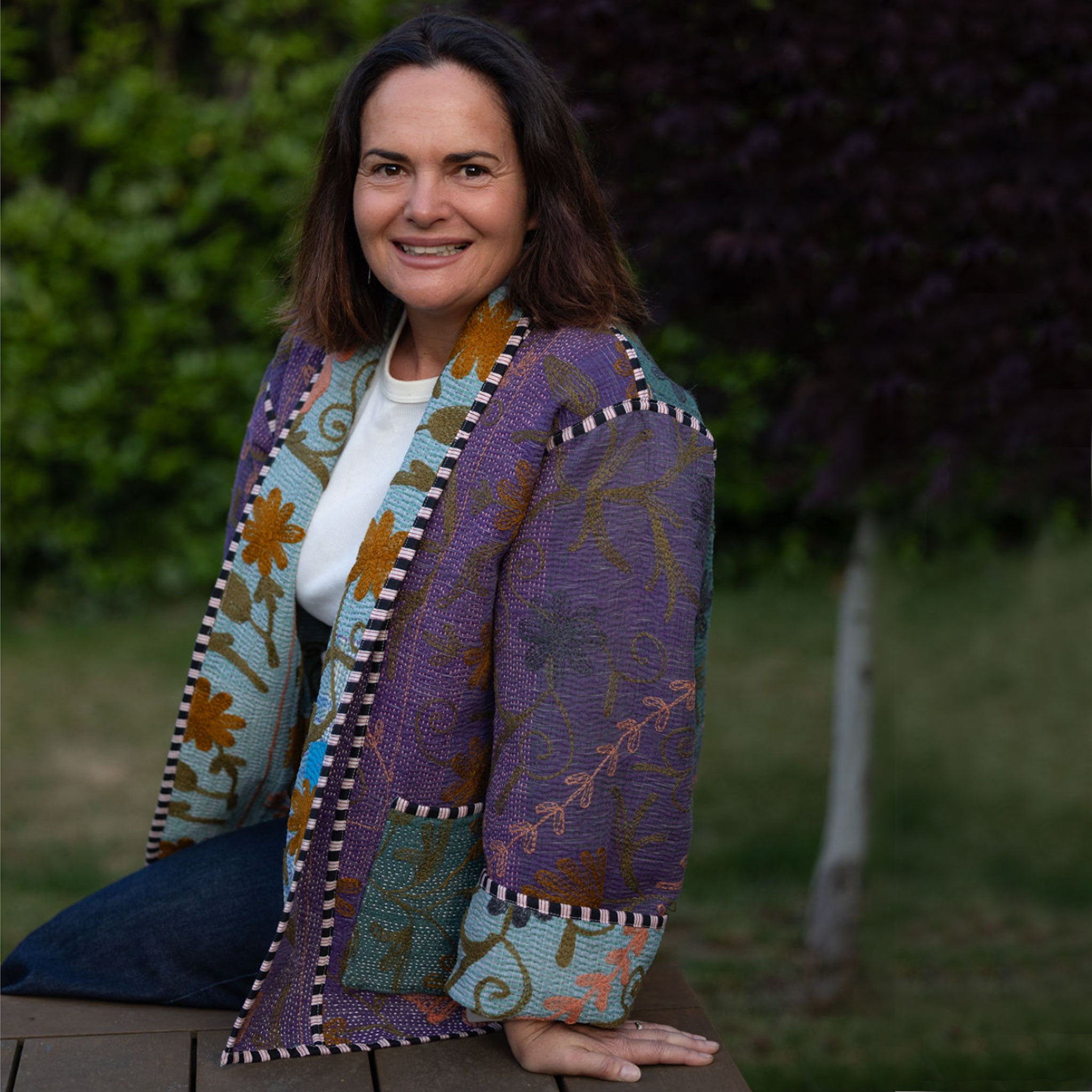 Woman (Tracey) wearing a colourful, patterned jacket sitting outdoors with greenery in the background